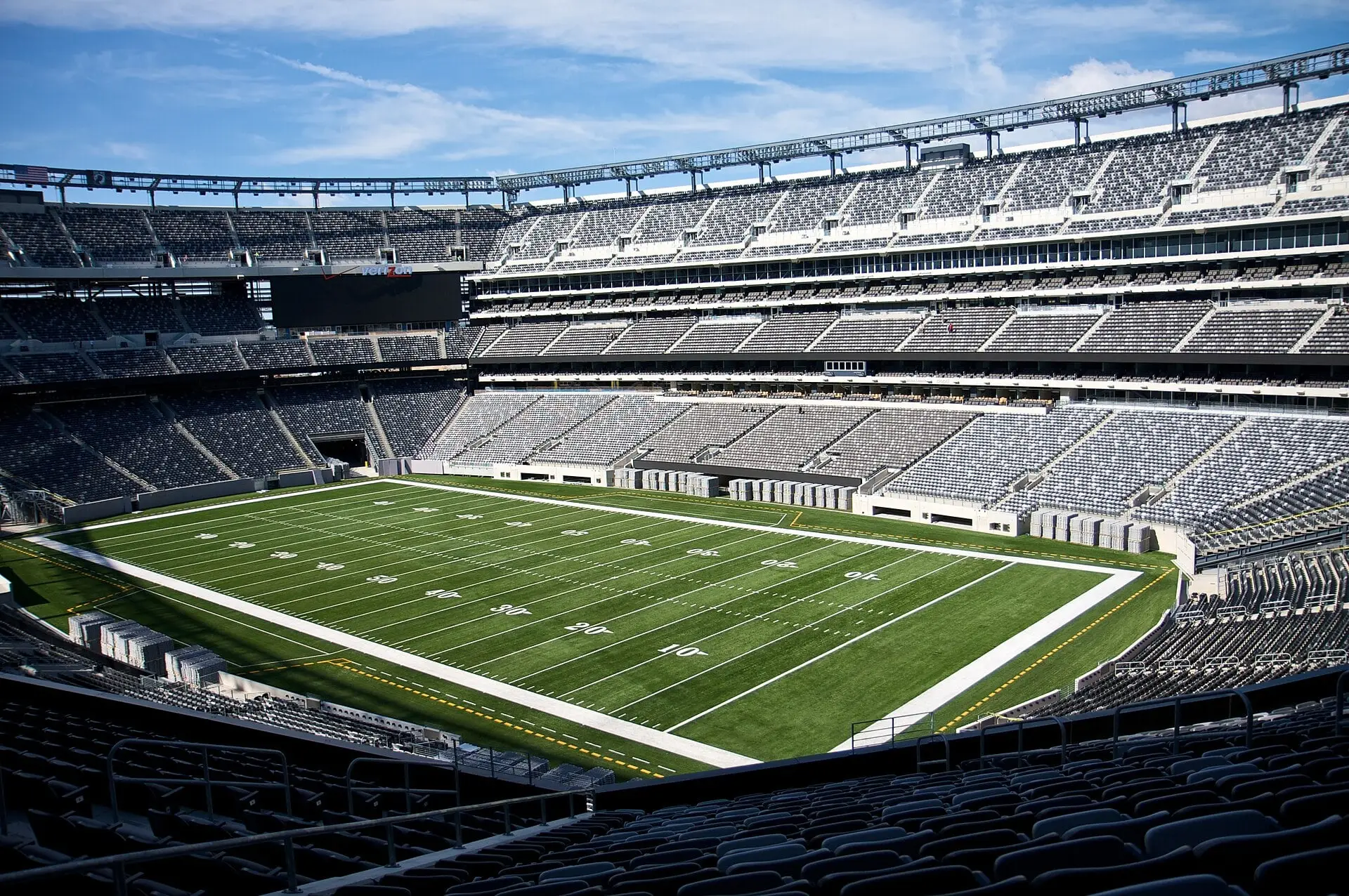 Empty MetLife Stadium seating bowl and field from the mezzanine corner under a blue sky, with the rim lighting truss above