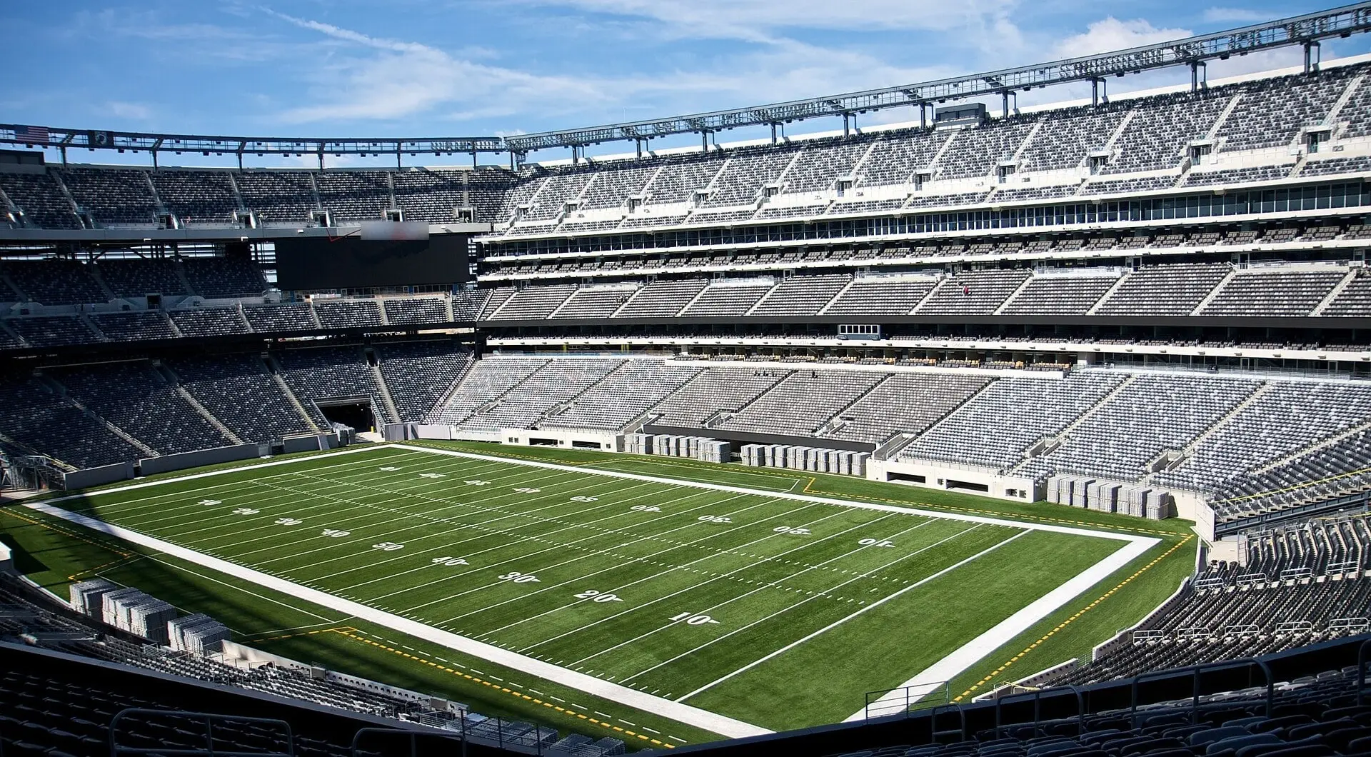 Empty MetLife Stadium seating bowl and field from the mezzanine corner under a blue sky, with the rim lighting truss above